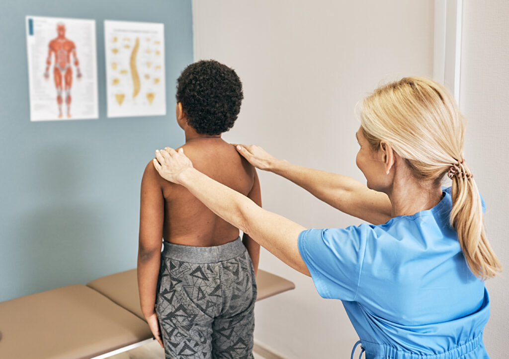 Healthcare provider examines a child's back and shoulder alignment during a physical assessment in an exam room.
