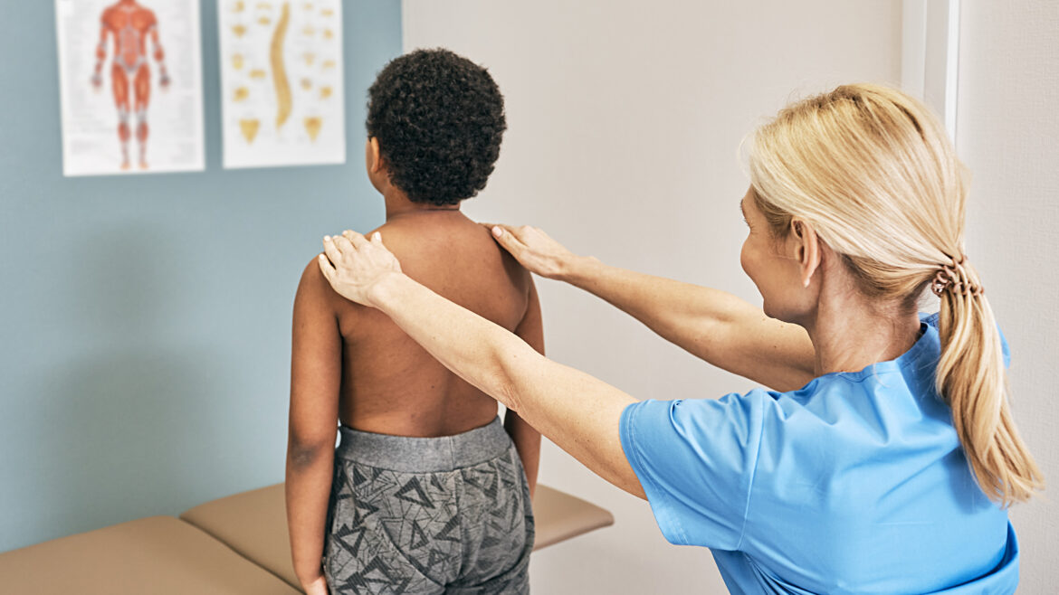 Healthcare provider examines a child's back and shoulder alignment during a physical assessment in an exam room.