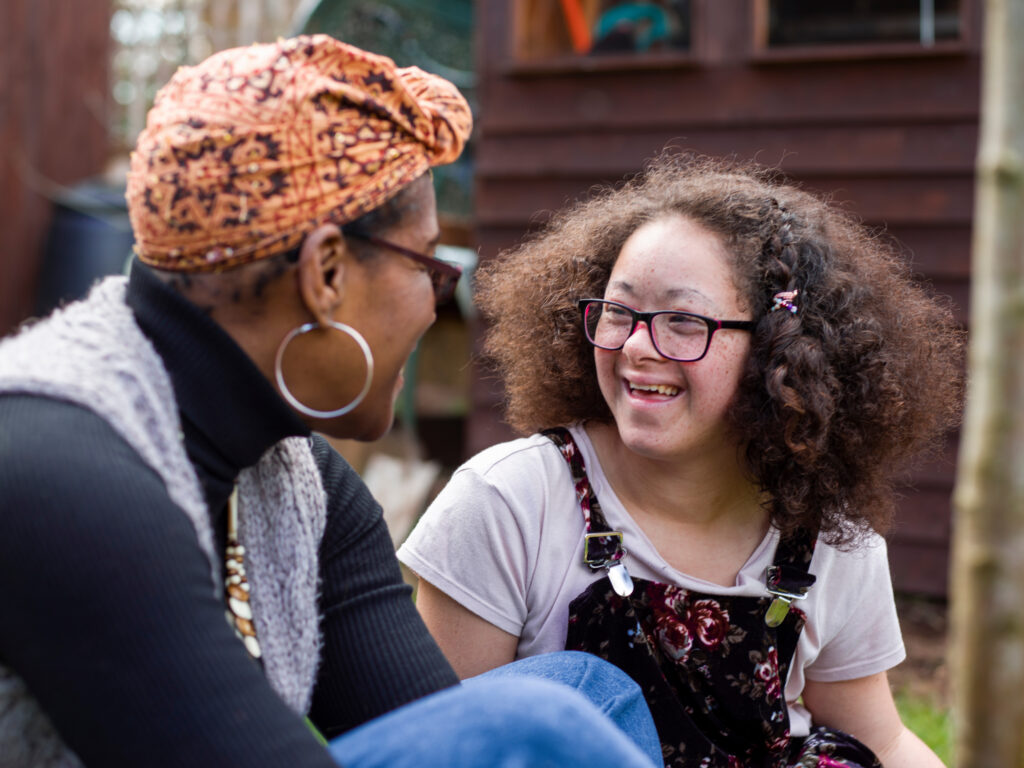 Outdoor photo of two people seated and facing one another in conversation. One person wears a patterned headscarf, large hoop earrings and a gray vest over a dark top. The other person has curly hair, glasses and floral-patterned overalls layered over a light shirt. Trees and a wooden structure are visible in the background.