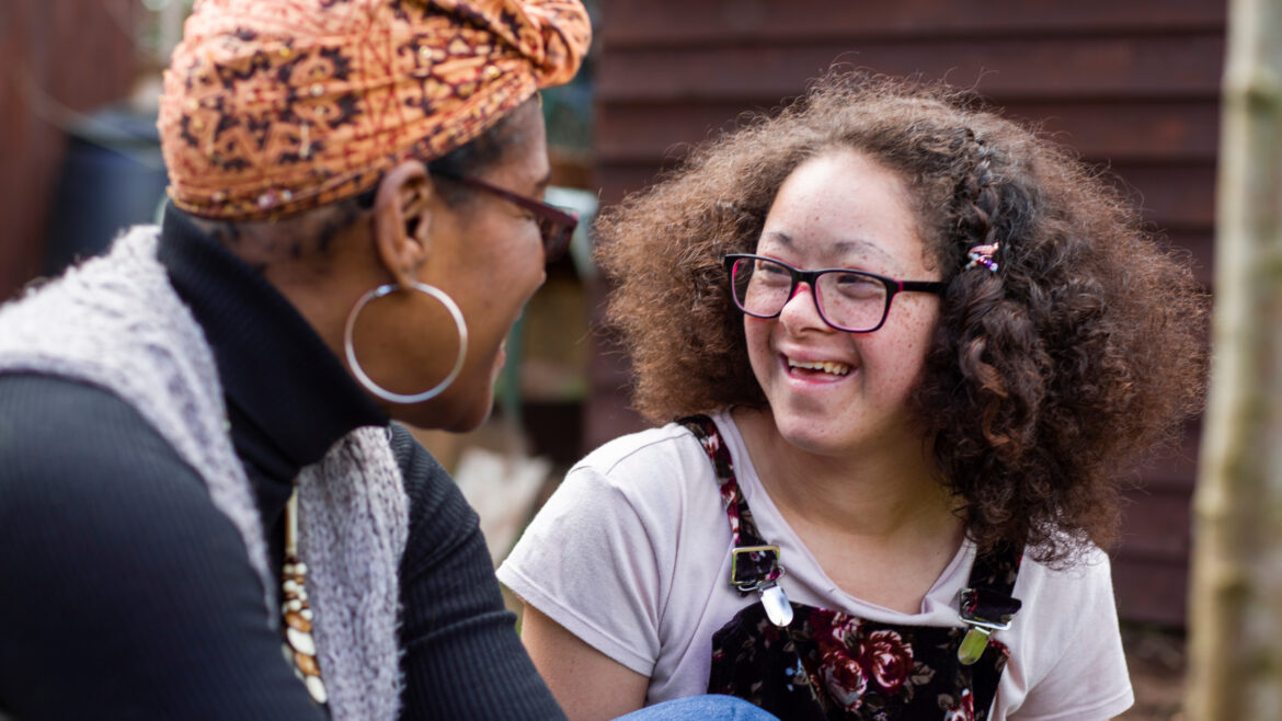 Outdoor photo of two people seated and facing one another in conversation. One person wears a patterned headscarf, large hoop earrings and a gray vest over a dark top. The other person has curly hair, glasses and floral-patterned overalls layered over a light shirt. Trees and a wooden structure are visible in the background.