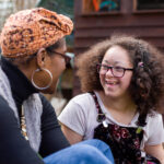 Outdoor photo of two people seated and facing one another in conversation. One person wears a patterned headscarf, large hoop earrings and a gray vest over a dark top. The other person has curly hair, glasses and floral-patterned overalls layered over a light shirt. Trees and a wooden structure are visible in the background.