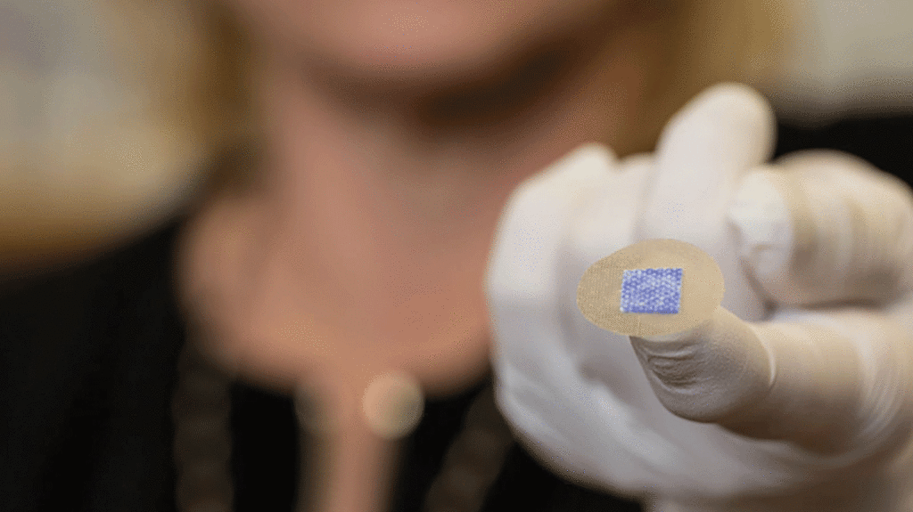 Close up, color image of someone extending a gloved hand with a small patch face up on their pointer finger that resembles a circular adhesive bandage that has a smaller, blue square patch on its center
