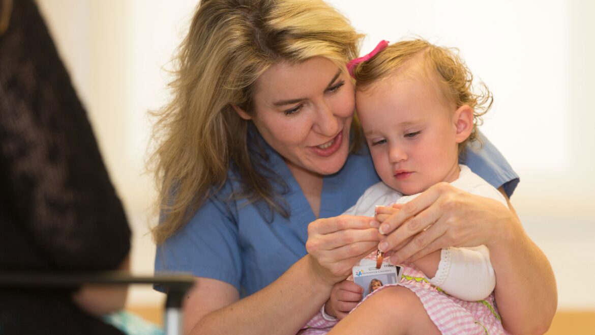 A healthcare provider engages warmly with a young child, helping her with a small medical item.