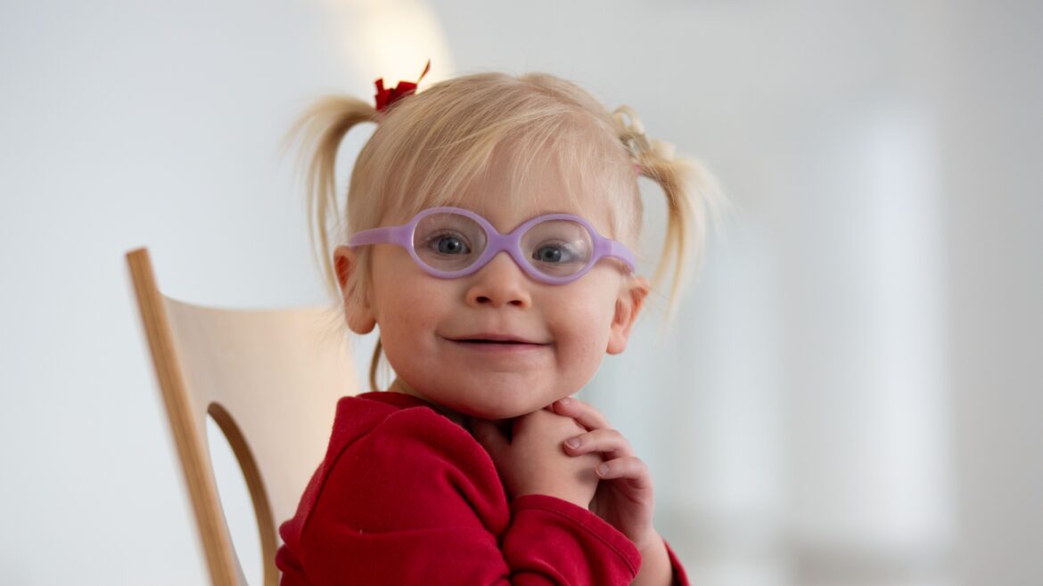 A young child in glasses smiles while sitting on a chair, suggesting a welcoming healthcare environment.