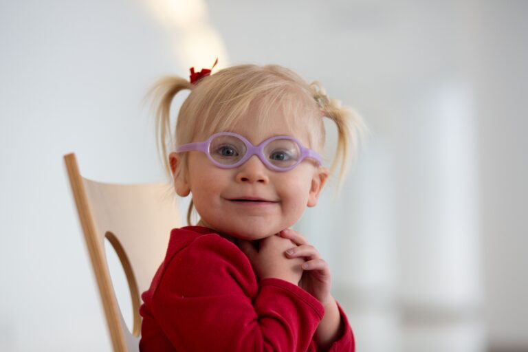 A young child in glasses smiles while sitting on a chair, suggesting a welcoming healthcare environment.