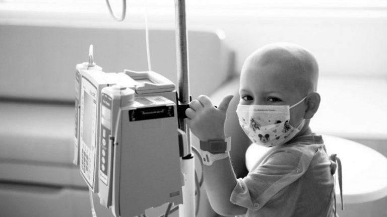 Black and white photo of a young patient with cancer wearing a mask and gown and holding an IV poll smiling and giving a thumbs up in the hospital
