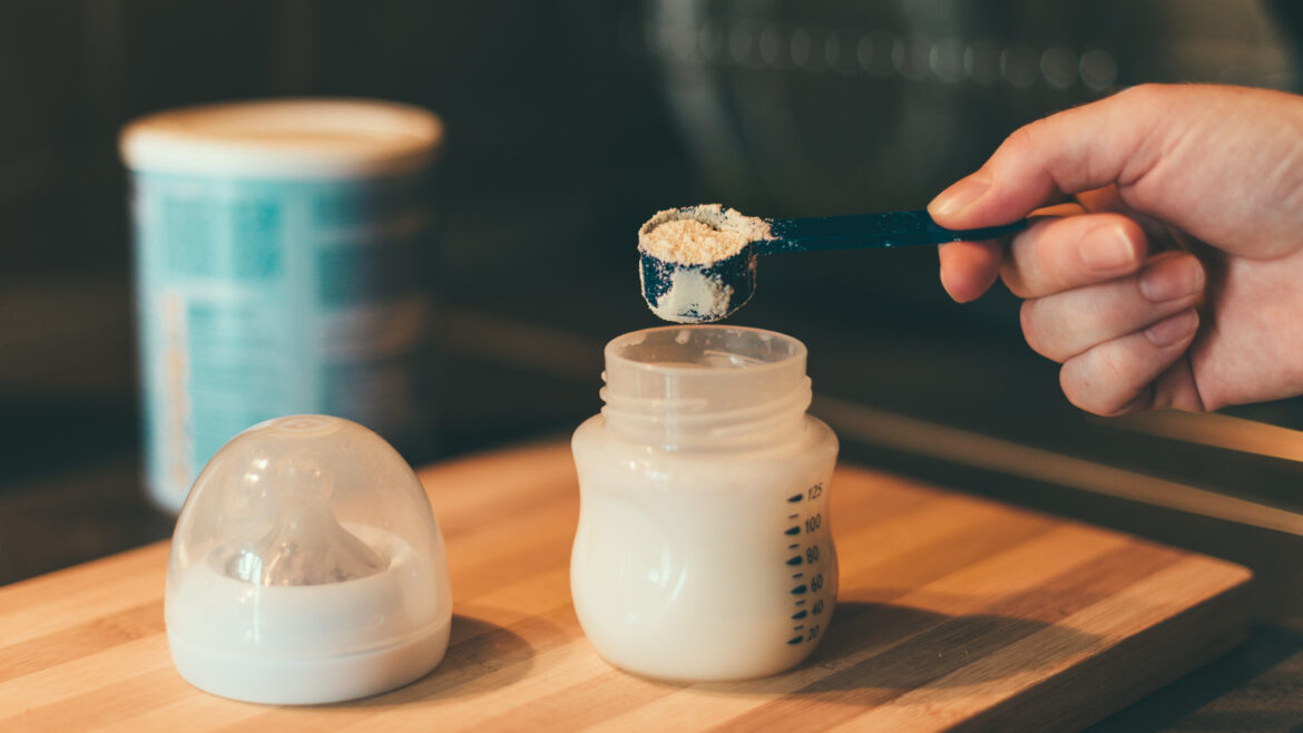 Image of a hand hovering above an infant formula bottle with a measuring spoon filled with powdered formula