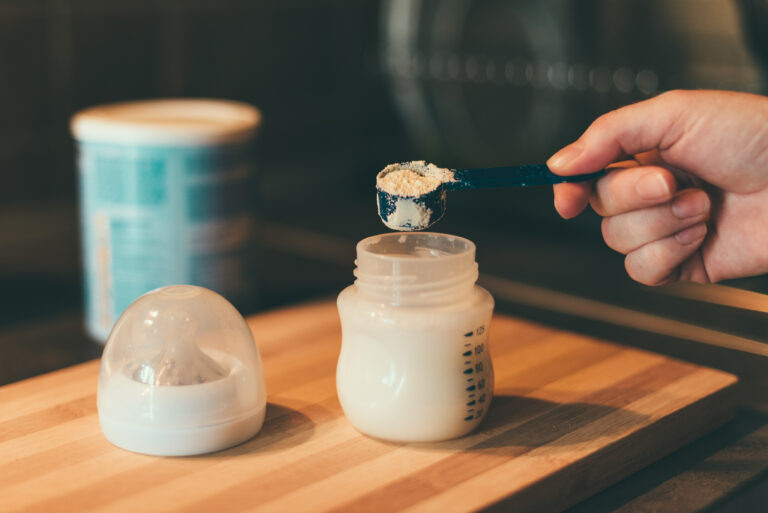 Image of a hand hovering above an infant formula bottle with a measuring spoon filled with powdered formula