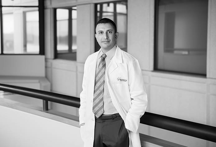 Black and white environmental portrait of Dr. Anup Patel standing in the hospital in his lab coat with his hand in his pocket