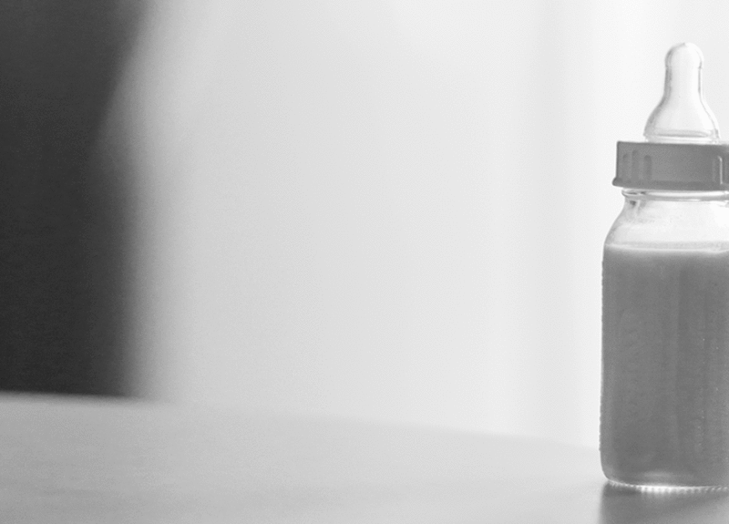 Black and white photo of baby bottle with formula on the table in front of a simple background