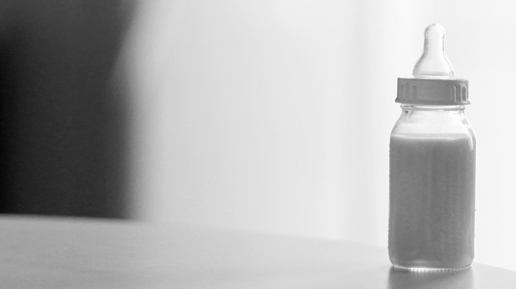 Black and white photo of baby bottle with formula on the table in front of a simple background