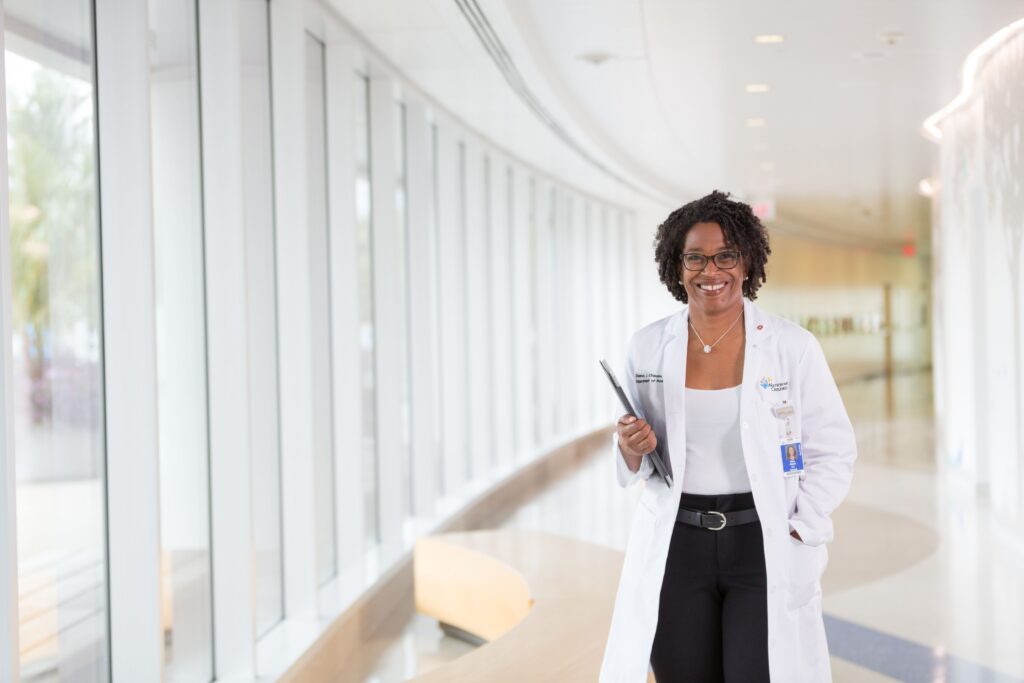 Deena Chisolm, PhD, smiles while walking down a brightly lit hospital corridor, holding a tablet and wearing a white coat.