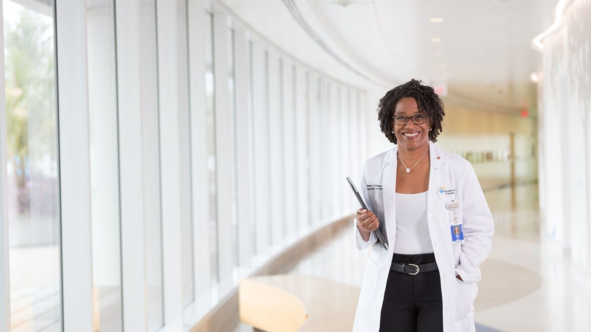 Deena Chisolm, PhD, smiles while walking down a brightly lit hospital corridor, holding a tablet and wearing a white coat.