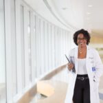 Deena Chisolm, PhD, smiles while walking down a brightly lit hospital corridor, holding a tablet and wearing a white coat.