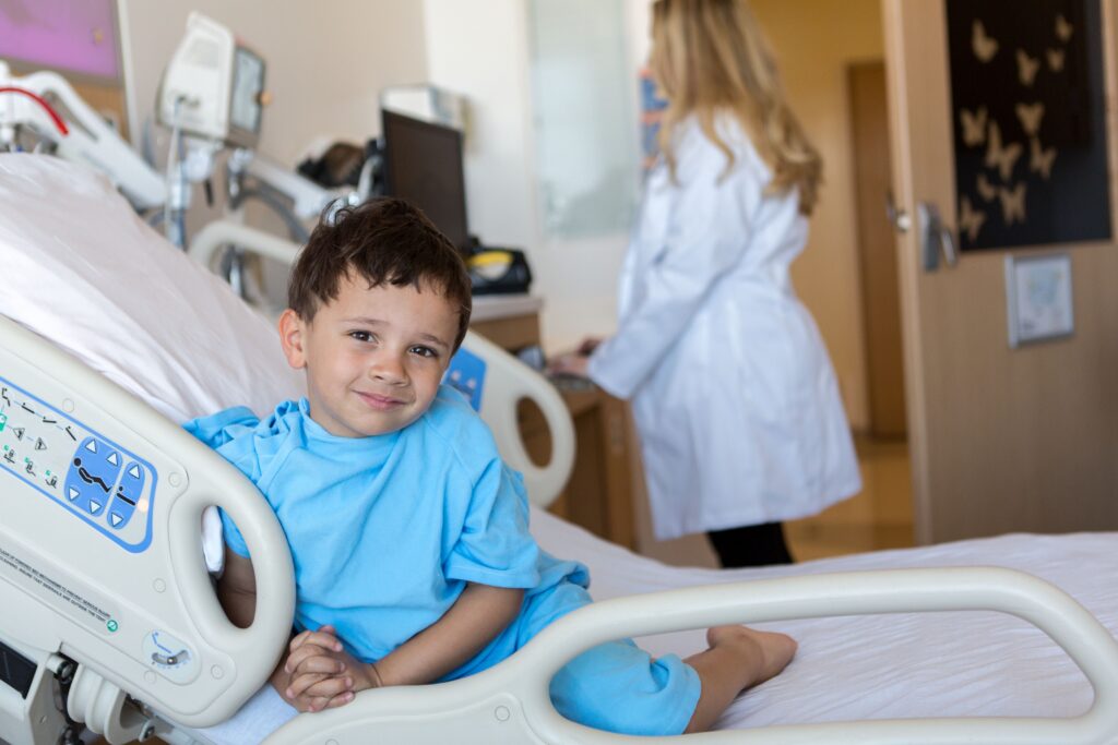 A young boy in a hospital bed smiles while wearing a blue hospital gown, with a healthcare professional in the background.