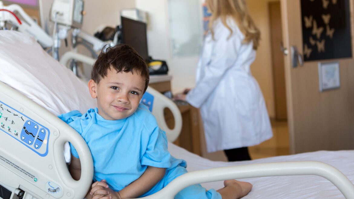 A young boy in a hospital bed smiles while wearing a blue hospital gown, with a healthcare professional in the background.