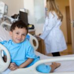 A young boy in a hospital bed smiles while wearing a blue hospital gown, with a healthcare professional in the background.