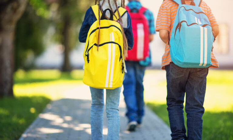 Three kids walking away with backpacks on a sunny day on a sidewalk