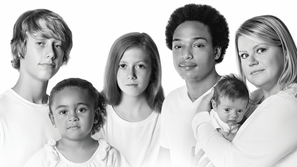 A black-and-white image of an adolescent White boy, a young Black girl, an adolescent White girl, a Black teen boy, and a young White mom and her infant, all in a row across the screen, all in white shirts, and all with solemn expressions.
