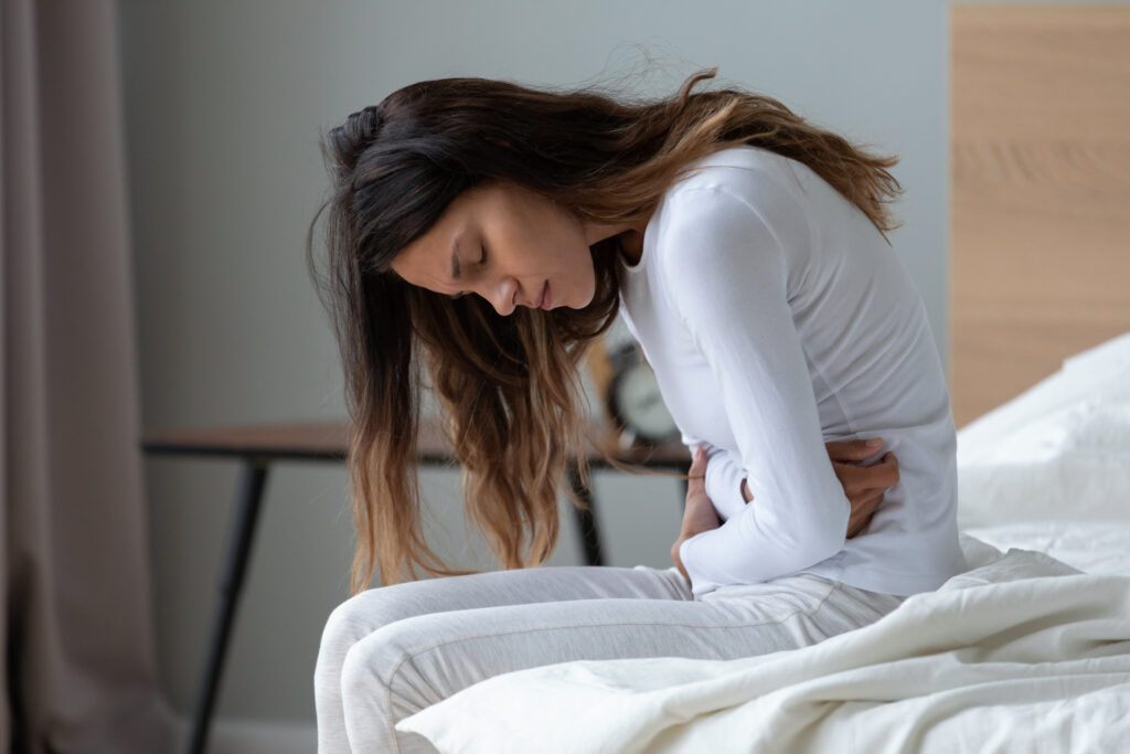 Sick young woman sit on bed at home touch belly suffer from stomach ache, stressed ill teen girl feel bad struggle with periods pain