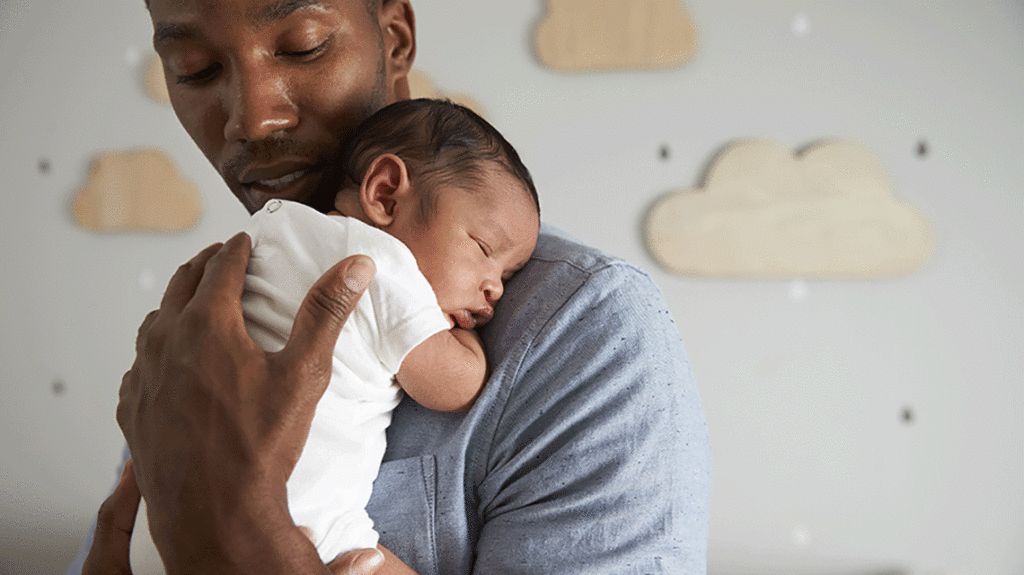 Color photo of Black father holding infant on shoulder in front of nursery background with clouds on the wall