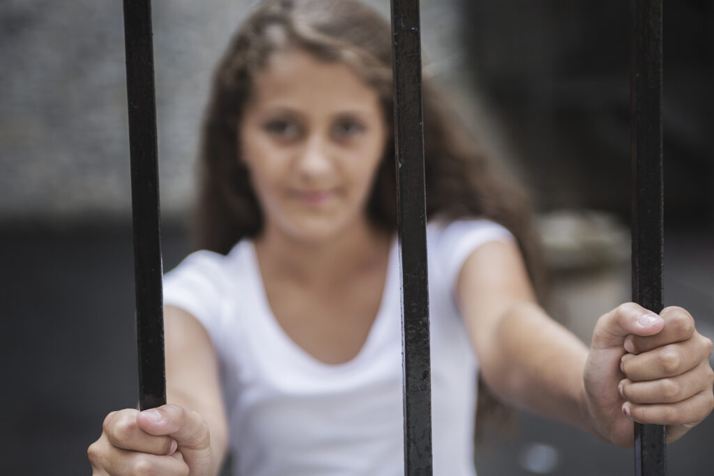 Young girl in a white tshirt is on the other side of a barred fence, face blurred and focus on her hands on the bars