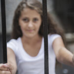Young girl in a white tshirt is on the other side of a barred fence, face blurred and focus on her hands on the bars