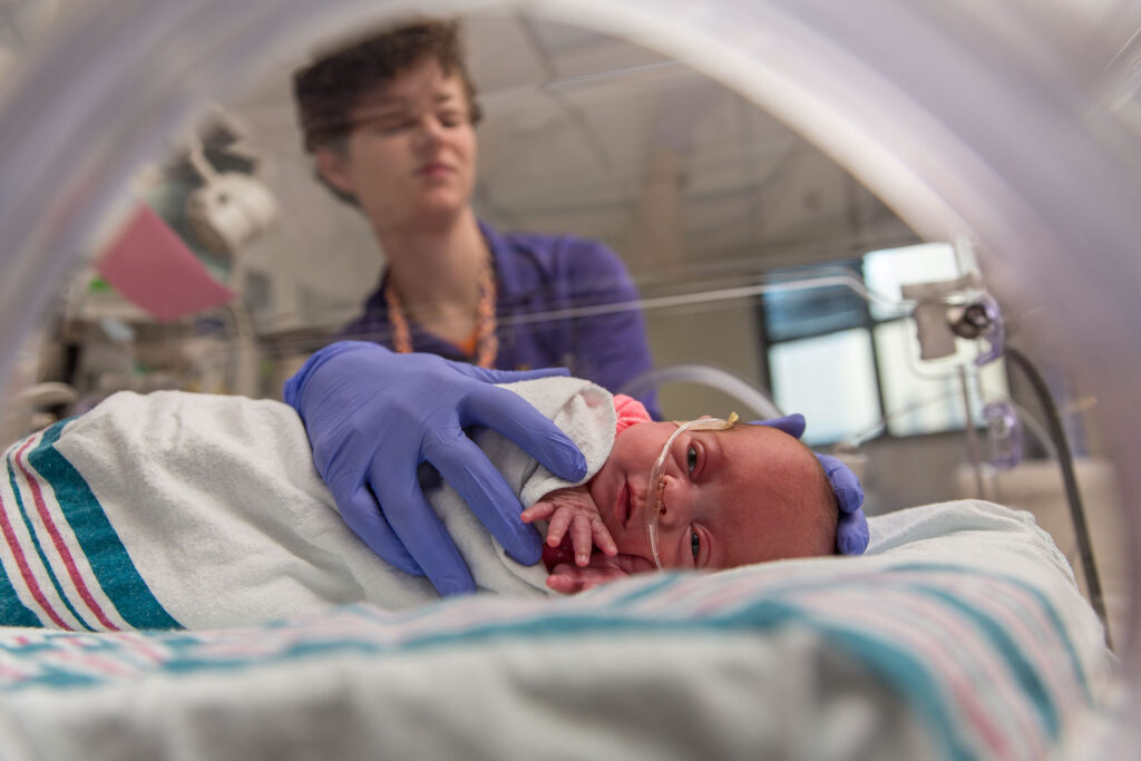 Premature baby receiving respiratory support through a nasal cannula in a neonatal intensive care unit, monitored and cared for by a health care provider.