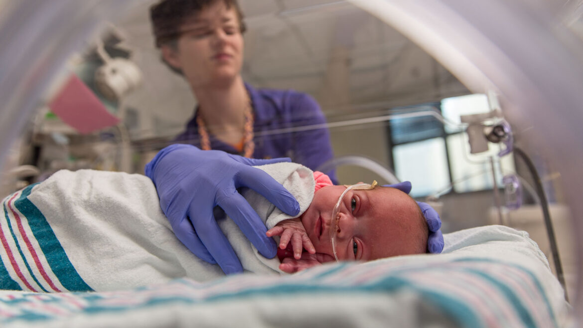Premature baby receiving respiratory support through a nasal cannula in a neonatal intensive care unit, monitored and cared for by a health care provider.