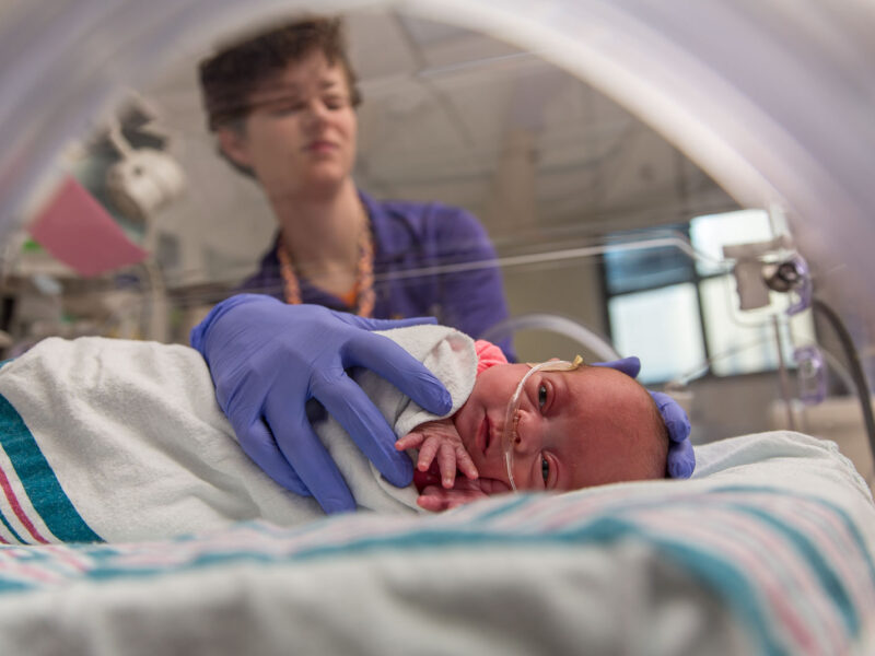 Premature baby receiving respiratory support through a nasal cannula in a neonatal intensive care unit, monitored and cared for by a health care provider.