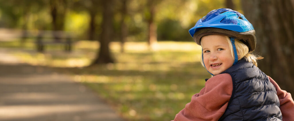Smiling child wearing a blue bike helmet and vest while riding outdoors on a sunny day, promoting bicycle safety and healthy outdoor activities for kids.