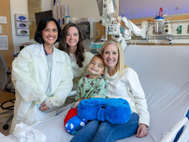 Mother and son sitting in hospital bed with doctors Allison Bradbury and Emily de los Reyes standing at the bedside. Everyone is excited; soon the child will receive a brand new gene therapy for his ultrarare disease.