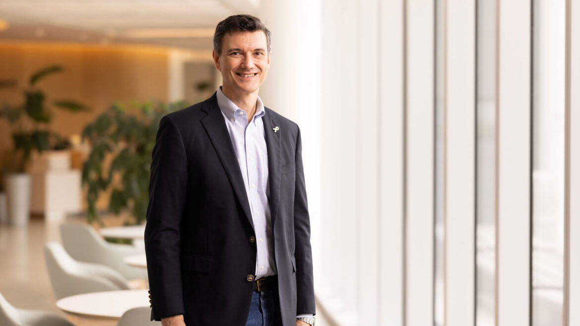 Portrait of Alexander Bishop, DPhil, standing in a hospital lobby and smiling, wearing a navy blazer and light blue shirt.