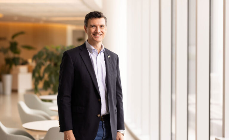 Portrait of Alexander Bishop, DPhil, standing in a hospital lobby and smiling, wearing a navy blazer and light blue shirt.