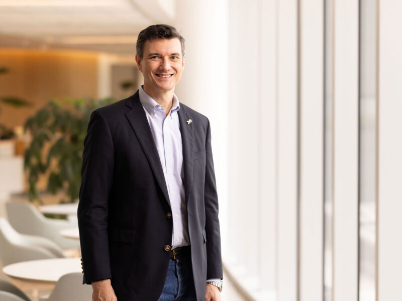 Portrait of Alexander Bishop, DPhil, standing in a hospital lobby and smiling, wearing a navy blazer and light blue shirt.