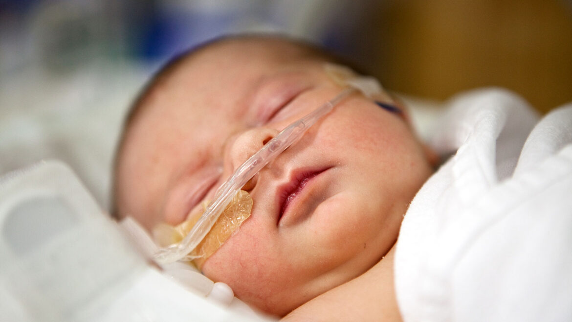 Newborn baby with nasal canula oxygen support receiving care in a neonatal intensive care unit (NICU) at a hospital, resting while monitored by medical staff.
