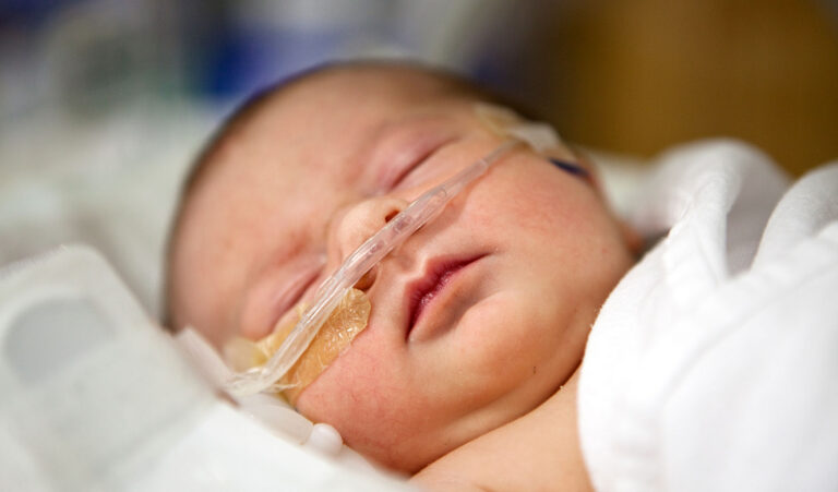 Newborn baby with nasal canula oxygen support receiving care in a neonatal intensive care unit (NICU) at a hospital, resting while monitored by medical staff.