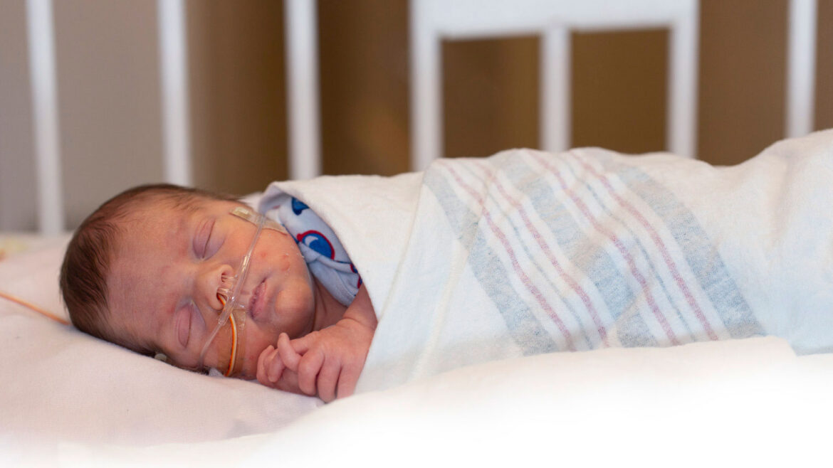 Newborn with bronchopulmonary dysplasia sleeping in a NICU crib, wrapped in a hospital blanket and receiving oxygen through a nasal cannula.