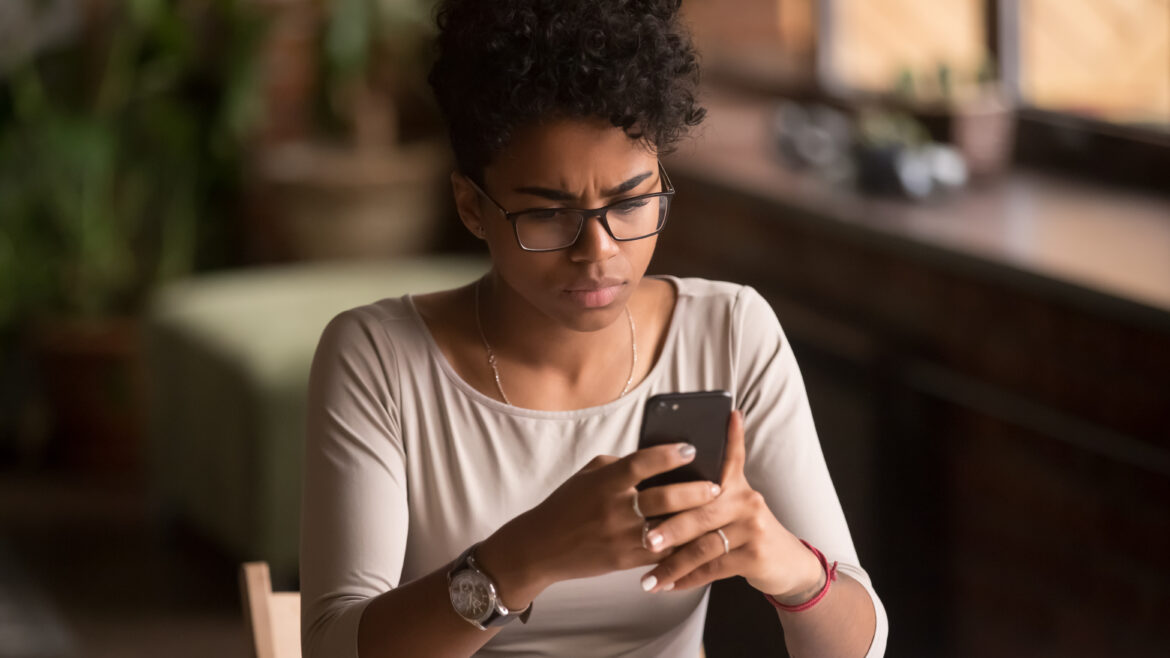 Upset confused Black woman holding cellphone having problem with mobile phone, frustrated angry mixed race girl reading bad news in message looking at smartphone annoyed by spam or missed call