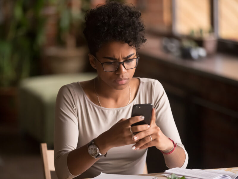 Upset confused Black woman holding cellphone having problem with mobile phone, frustrated angry mixed race girl reading bad news in message looking at smartphone annoyed by spam or missed call