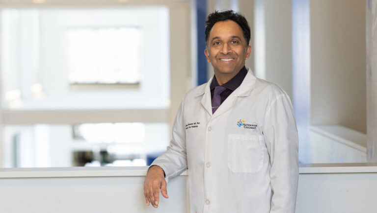 Ashley Fernandes, MD, PhD, wearing a white coat with a Nationwide Children's logo, standing in front of a blurred hospital interior background