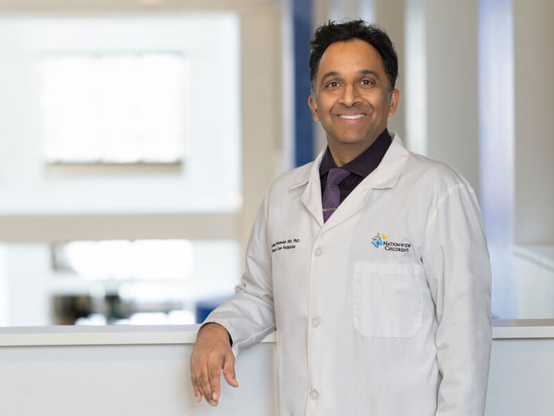 Ashley Fernandes, MD, PhD, wearing a white coat with a Nationwide Children's logo, standing in front of a blurred hospital interior background