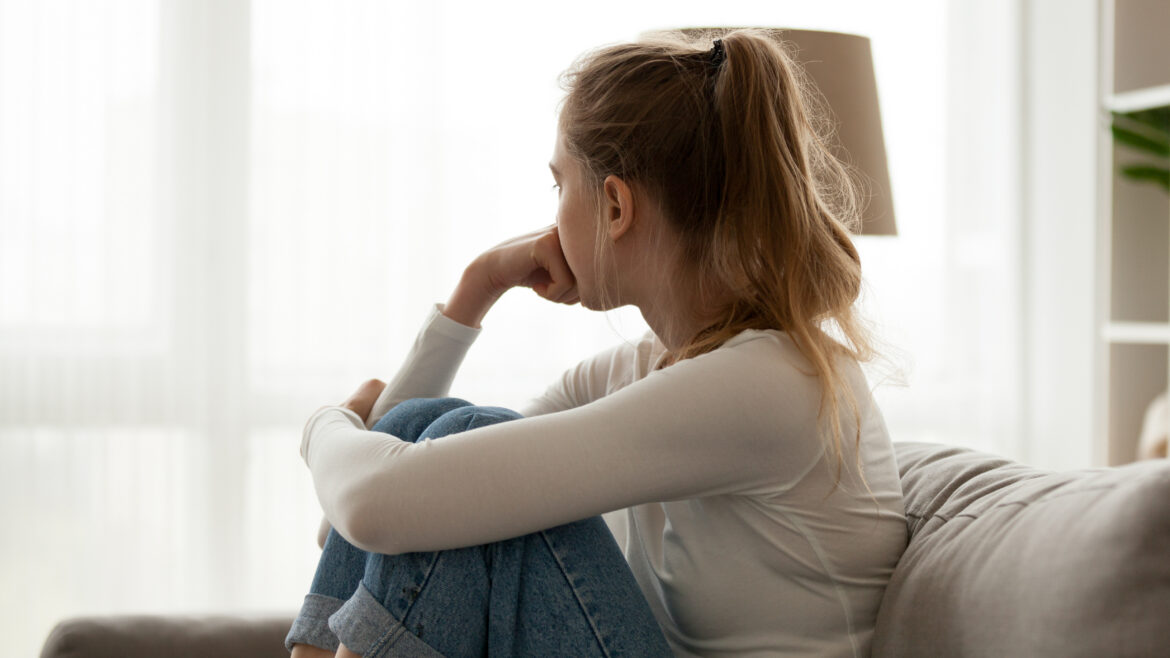 Teen sitting curled up on a couch, looking out a bright window, appearing thoughtful or pensive.