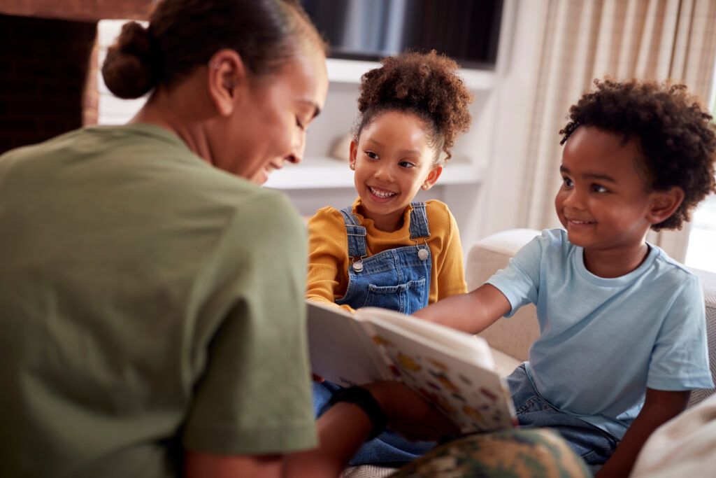 A woman sitting on a couch reading a book with two young children, who smile and look at the pages together.