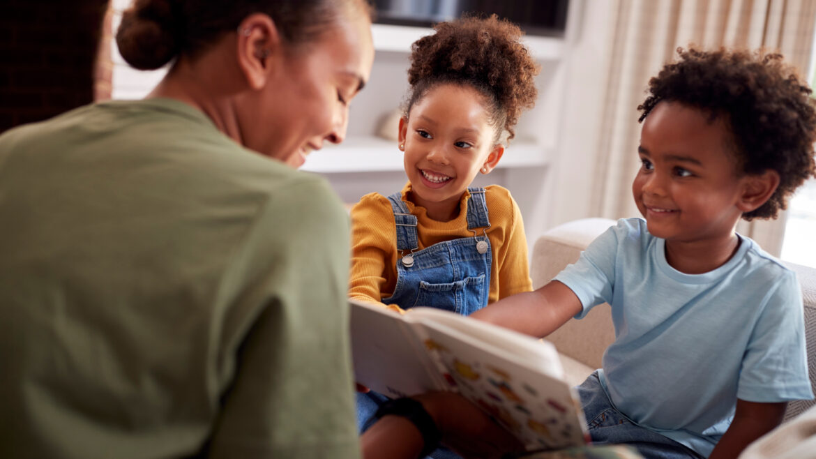 A woman sitting on a couch reading a book with two young children, who smile and look at the pages together.