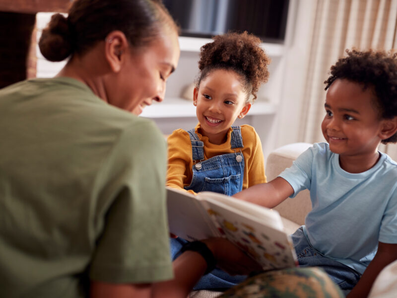 A woman sitting on a couch reading a book with two young children, who smile and look at the pages together.