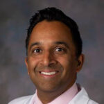 Studio portrait of Ashley Fernandes, MD, PhD, he is wearing a white coat, pink shirt and tie.
