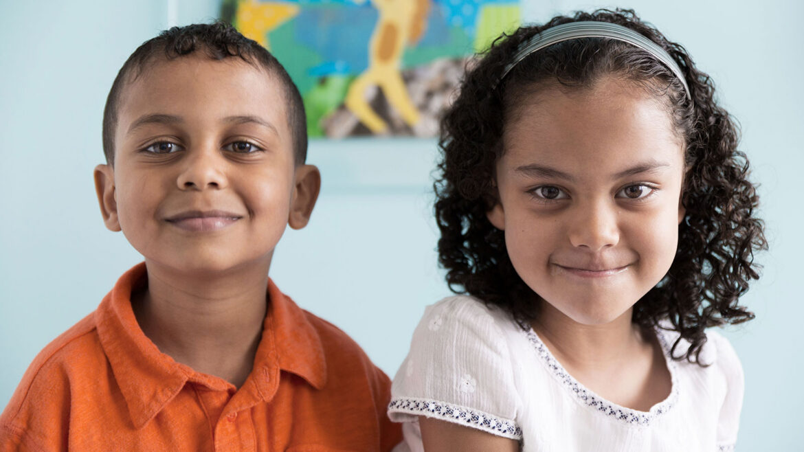 Two smiling young children sit side by side indoors, facing the camera, with a colorful blurred artwork in the background.