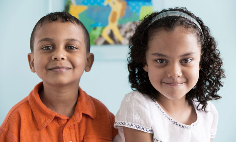 Two smiling young children sit side by side indoors, facing the camera, with a colorful blurred artwork in the background.