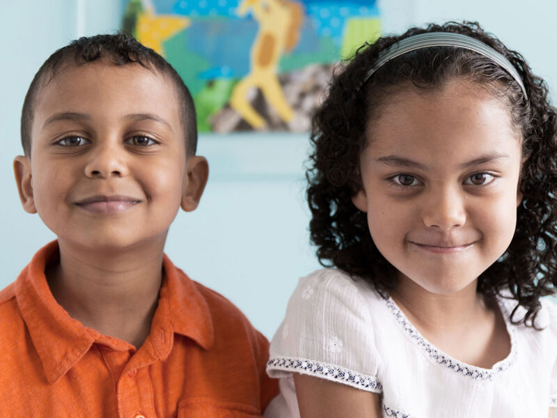 Two smiling young children sit side by side indoors, facing the camera, with a colorful blurred artwork in the background.